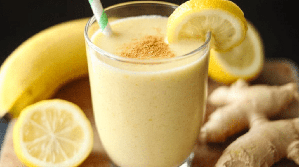 Creamy Banana Lemon Ginger Smoothie in a glass with a green and white striped straw, garnished with a lemon slice and cinnamon sprinkle, surrounded by fresh banana, lemon slices, and ginger root on a wooden surface.