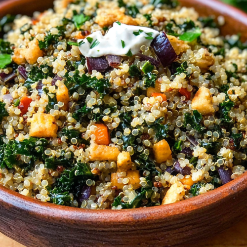 A wooden bowl filled with quinoa kale pilaf containing dark leafy greens, golden quinoa grains, orange carrots, purple onions, and tofu cubes, topped with a small dollop of white sauce.