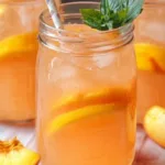 Glass mason jar of homemade peach lemonade with fresh peach slices, mint garnish, and ice, alongside fresh peach wedges and a pitcher of lemonade in the background